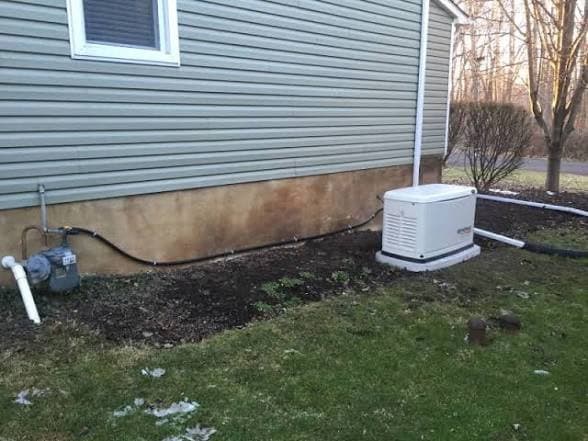 Standby generator installed on a gravel pad with gas line and conduit run along gray vinyl siding.
