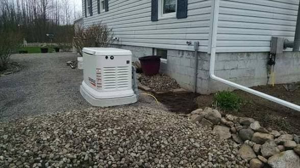 Standby generator installed on a gravel pad near a stone landscaping bed beside a gray-sided home.
